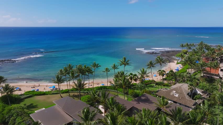 Aerial pan of Poipu Beach on Kauai Island, Hawaii, showing turquoise waters, sandy shoreline, swaying palm trees, and nearby luxury homes.