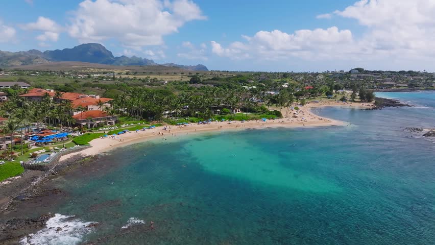 Sweeping aerial pan of Poipu Beach on Kauai Island, Hawaii, showing golden sand, turquoise waters, beachgoers, palm trees, and distant mountains.