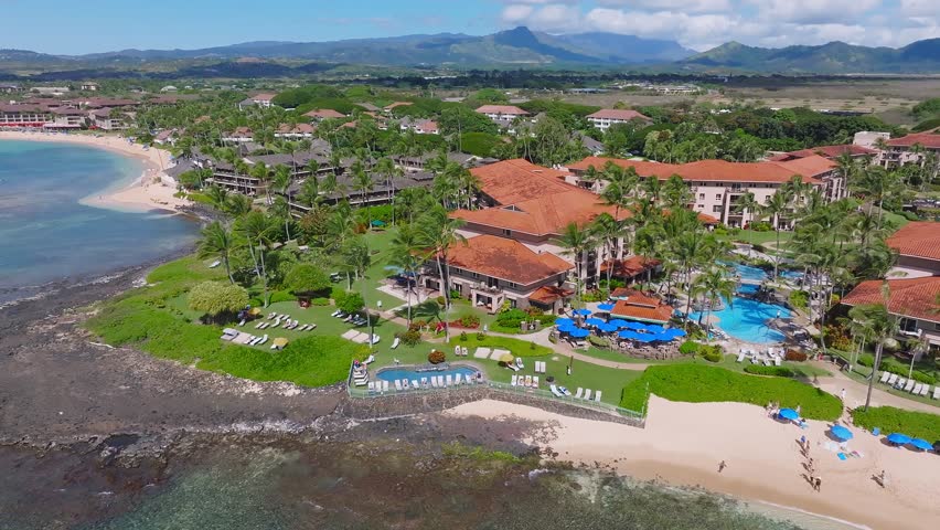 Aerial view of Poipu Beach on Kauai Island, Hawaii, showing turquoise waters, rocky shoreline, sandy beach, palm trees, and luxury resorts.