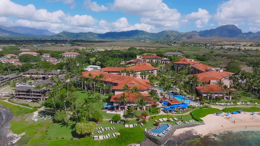 Aerial view of Poipu Beach on Kauai Island, Hawaii, showing turquoise waters, sandy shores, red roofed resorts, lush greenery, and rugged mountains.