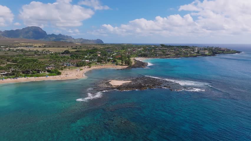 Aerial view of Poipu Beach on Kauai Island, Hawaii, showing turquoise waters, sandy shoreline, red roofed buildings, and rolling hills in the distance.
