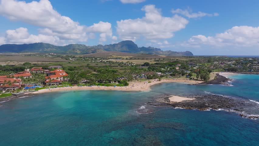 Sweeping aerial view of Poipu Beach with turquoise waters, sandy shores, rocky outcrops, red roofed buildings, and lush greenery under a blue sky.