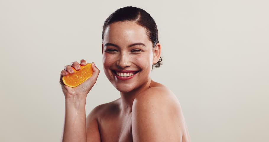 Woman, skincare and squeeze orange at face with smile, natural treatment or vitamin C in studio. Girl, citrus fruit or excited on white background space for organic care, healthy skin or antioxidants