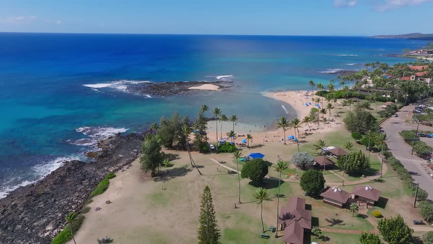 Aerial pan of Poipu Beach on Kauai Island, Hawaii, showing turquoise waters, sandy shoreline, palm trees, visitors, and nearby park areas.