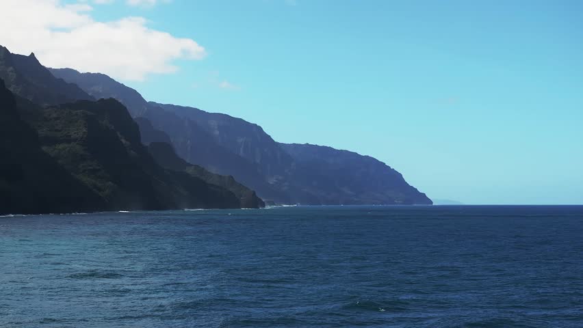 Aerial view of the Napali Coast on Kauai Island, Hawaii, with jagged cliffs, lush greenery, turquoise waters, and gentle ocean waves in motion.