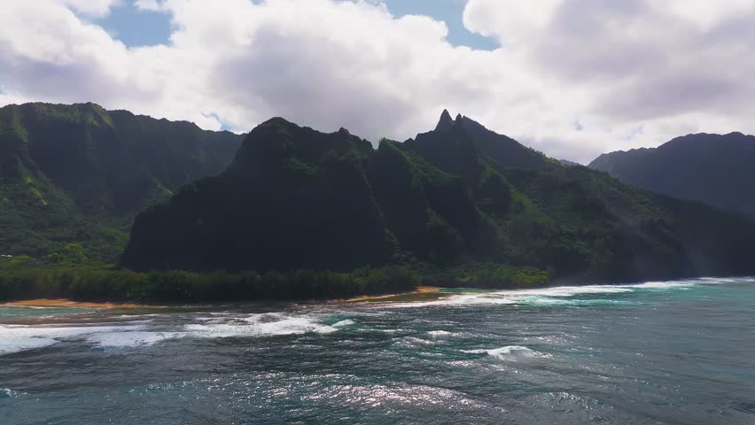 Aerial view of Napali Coast on Kauai Island, Hawaii, with jagged green cliffs, turquoise waters with whitecaps, and a narrow golden beach below.