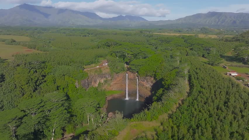 Aerial view of Wailua Falls on Kauai Island, Hawaii, with twin waterfalls flowing into a pool, surrounded by greenery, fields, and mountains.