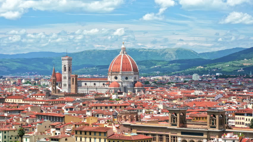 Panoramic view of Florence (Firenze), Italy, showcasing the city center with the Cathedral of Santa Maria del Fiore. Blue sky with clouds, rolling hills, and mountains in the background.