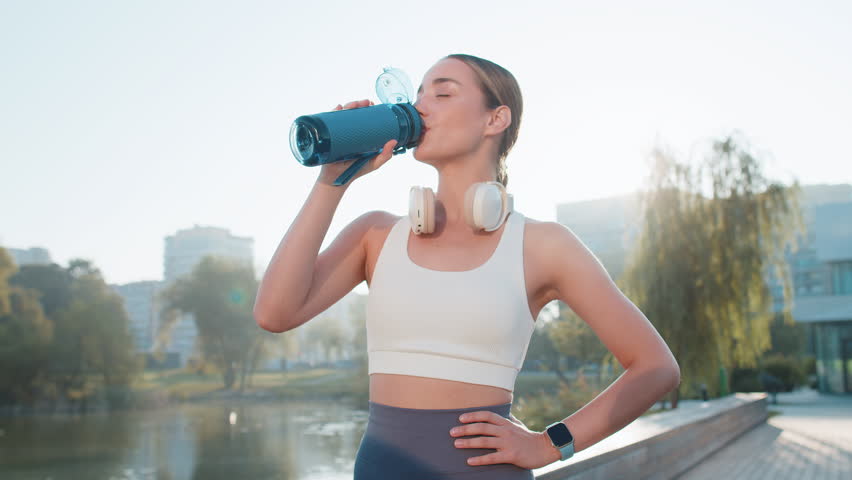 Athletic fit sport runner girl drinking water from bottle after training exercise. Workout cardio in park in morning. Young slim woman jogger enjoy healthy lifestyle. Active sportswoman activities.