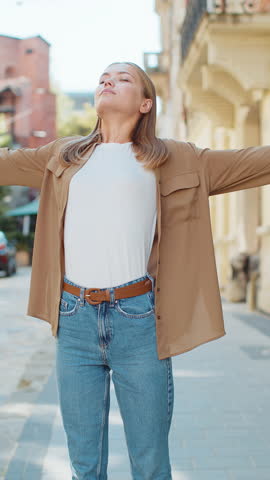 Caucasian young woman taking a deep breath of fresh air, relaxing, taking a break, resting, meditating, calm down outdoors. Girl enjoying happy day standing in urban city street. Town lifestyles.