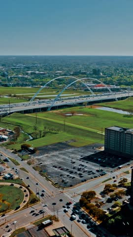 This aerial shot captures a vast expanse of Dallas, highlighting a prominent arch bridge over a busy highway amidst green fields and urban structures.