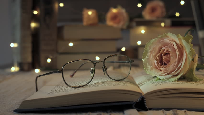 A tranquil reading nook features an open book resting on a table, accompanied by stylish glasses and a delicate pink rose, all softly illuminated by fairy lights creating a warm atmosphere.