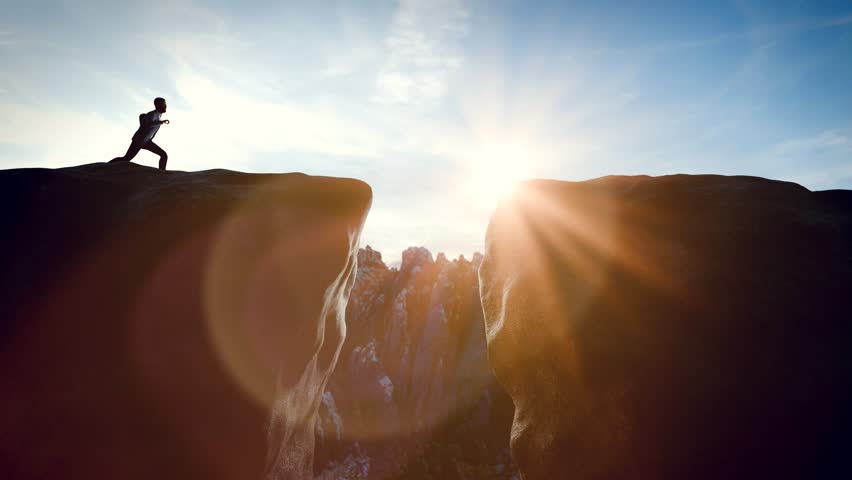 Courageous man jumping over a gap on a mountain peak at sunrise.