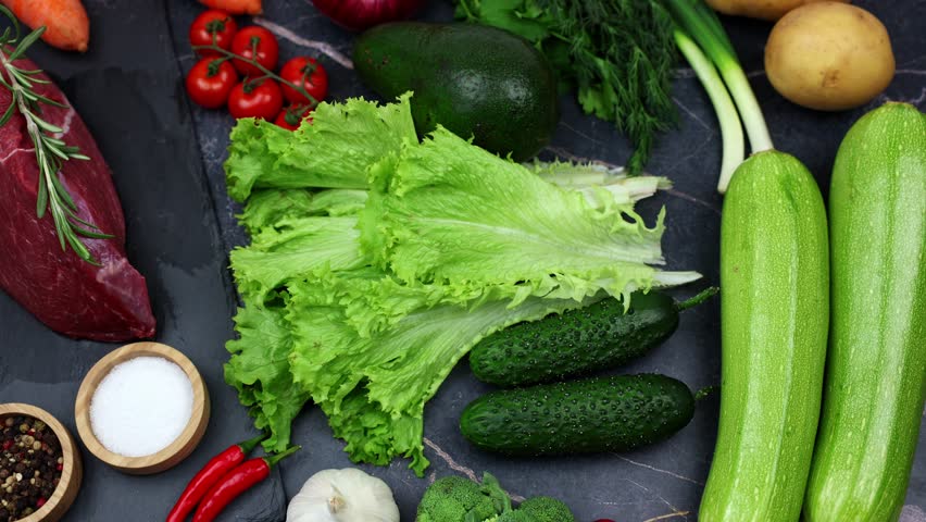 Top view of a piece of beef with a sprig of rosemary on top, vegetables, herbs and seasonings lying nearby on a dark background. The concept of proper nutrition, natural products, the cooking process