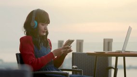 Stylish asian woman listening music in headphones on mobile phone app after video conference meeting with colleagues, online working on laptop. Beautiful twilight scene background. - Powered by Shutterstock - Get 15% off with code: PIKWIZARD15