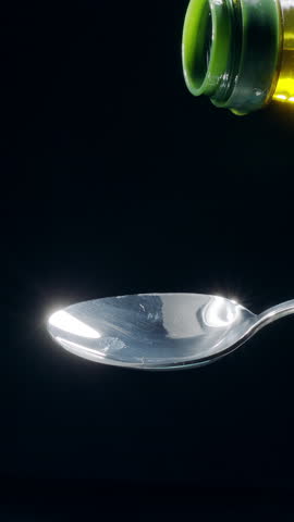 Sparkling golden olive oil is being poured into a spoon and overflowing it on a black background. Slow motion vertical template.