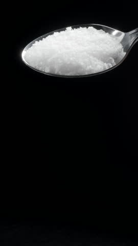 Adding salt or sprinkling salt. Salt is falling down from a silver spoon black background. Slow motion vertical template. 