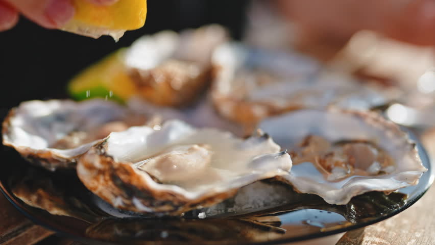 Woman hand pouring oysters juice squeezing lemon preparing to eat in restaurant outdoors in sunny day, close-up view. Edible fresh raw delicacy seafood. Mediterranean cuisine culinary food concept.