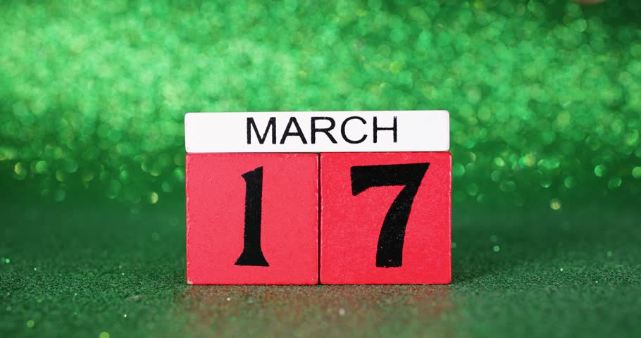 Footage of a red calendar block being placed down and picked back up again showing the date March the 17th which is the Irish St Patricks day for celebrations on a green glittery background