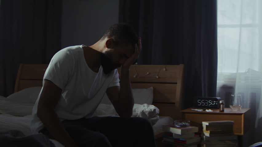 Young bearded man feels sorrow and pain sitting on messy bed. Male person deals with depression disorder walking through complicated life stage