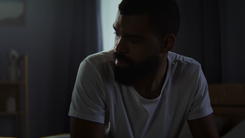 Bearded man leans onto fist feeling insecure at home. African American male seeks will to live after traumatic event in untidy bedroom. Mental health