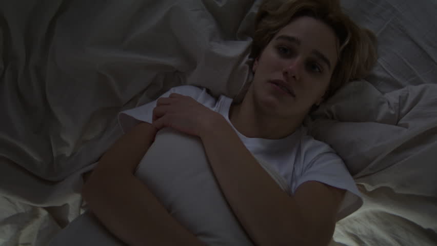 Upset young woman hugs pillow lying in bed at night. Blonde female with dark circles under eyes due to crying tries to deal with negative emotions