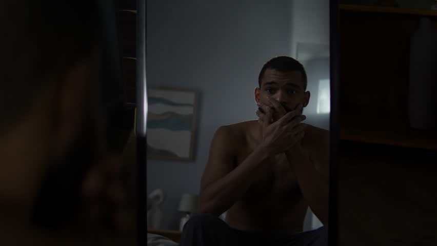 Pensive shirtless black man stares into mirror sitting in bedroom. African American guy feels life uncertainties reflecting emotional struggle