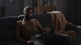 African American man smokes cigarette sitting shirtless on sofa. Depressed black man evokes sense of solitude and introspection in home setting - Powered by Shutterstock - Get 15% off with code: PIKWIZARD15