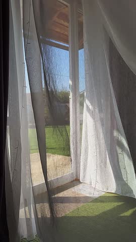 Room interior with open window overlooking back yard with lawn. Transparent tulle with morning sun rays on shadow background. Vertical video. High quality footage