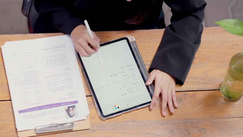 A woman is writing on a tablet with a pen. She is sitting at a desk with a clipboard and a vase - Powered by Shutterstock - Get 15% off with code: PIKWIZARD15