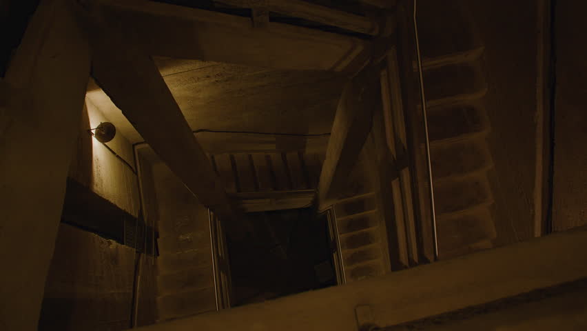 A dimly lit concrete stairwell with sharp angles, steel railings, and exposed structural beams, spiraling downward into darkness, creating a claustrophobic and industrial atmosphere.