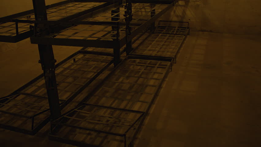 Tilt shot of old bunk beds with wooden planks on top, suspended by chains. The room has aged walls, exposed piping, and a ceiling lamp casting a warm glow. Bunk room in Fort de Mutzig fortress.