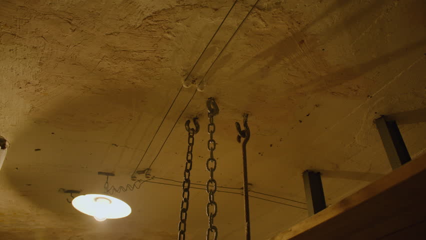 Tilt shot of a dimly lit bunk room with bunk beds suspended by chains. The room has aged walls, exposed piping, and a ceiling lamp casting a warm glow. Bunk room in Fort de Mutzig fortress.