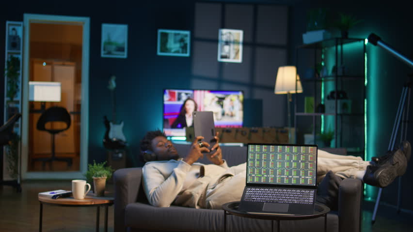 Black guy listens to music and reads from tablet on the couch, waiting for stock market trends to grow and increase his income. Young adult follows financial figures on laptop, investment. Camera B.