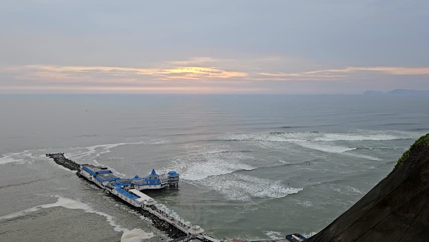 Pier and surfers sitting in rolling waves during a sunset in Miraflores district of Lima, Peru. Lima is the capital and the largest city of Peru.