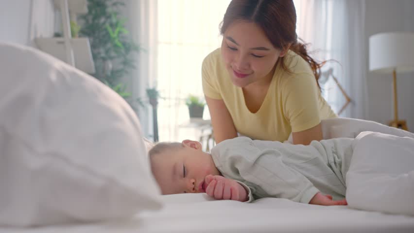Asian mother kissing her baby boy while sleep on bed in bedroom at home. Attractive parent look after young infant toddler lying down and resting comfortably, sleep for early childhood development.