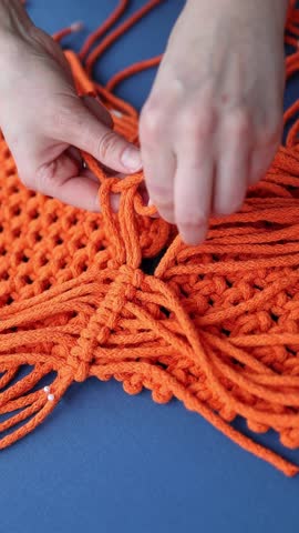 Female hands weaving macrame bag with orange ropes over blue background. Handmade decor piece process in slow motion. Intricate knot patterns. Bohemian charm in interior spaces, vertical shot