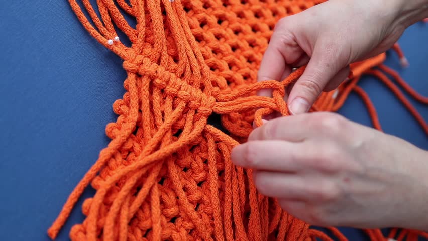 Female hands weaving macrame bag with orange ropes over blue background. Handmade decor piece process in slow motion. Intricate knot patterns. Bohemian charm in interior spaces
