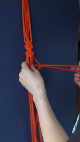 Female hands weaving macrame bag with orange ropes over blue background. Handmade decor piece process in slow motion. Intricate knot patterns. Bohemian charm in interior spaces, vertical shot
