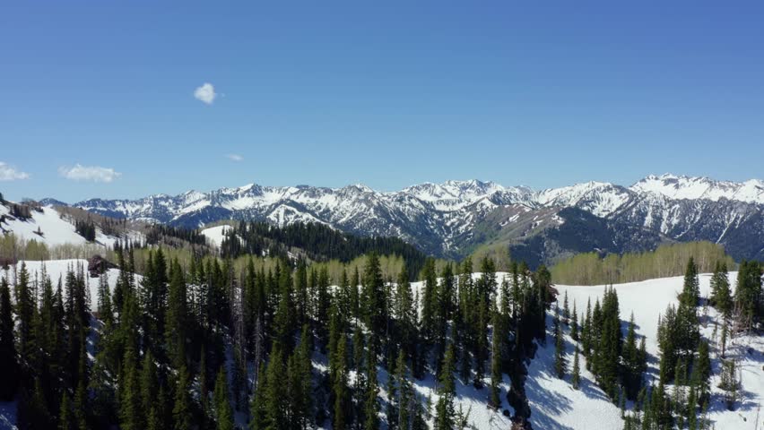 Wide landscape aerial drone shot of the Rocky Wasatch Mountains in Utah’s Big Cottonwood Canyon. The camera trucks right showing patchy summer snow among pine trees surrounded by rugged terrain
