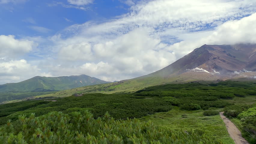 A majestic mountain, ASAHI-DAKE: The heart of Daisetsuzan National Park, rises above a lush green landscape under a bright blue sky with scattered clouds. A winding dirt road leads through the verdant