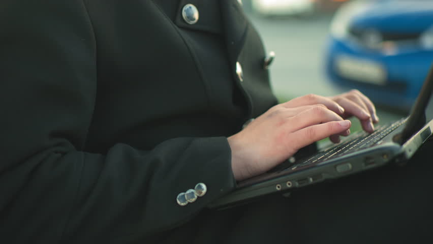 Close up of hands typing on laptop outdoors while wearing black suit with silver buttons, against blurred background featuring parked cars and greenery