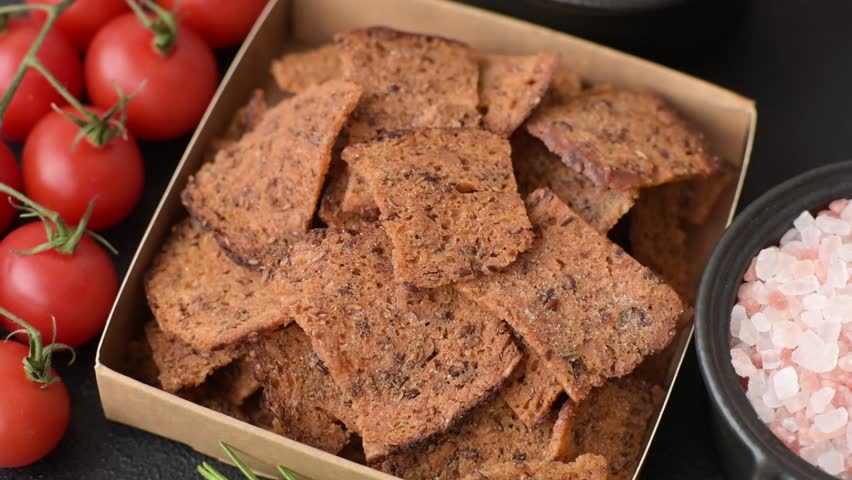 Crispy homemade brown bread chips with seeds and grains, salt, spices and herbs on a dark concrete background
