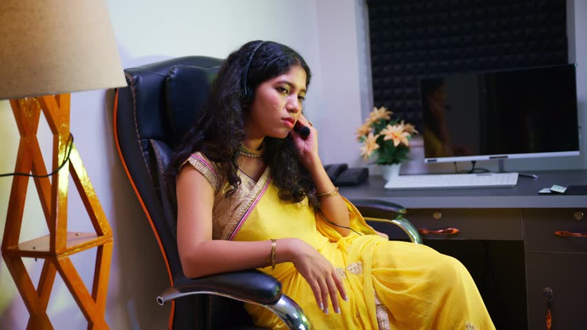 Young woman seated in call center speaks into a headset, Lady wears formal attire talk on microphone at desk, Girl sitting by her workstation, Female handles customer queries over phone, Communicates
