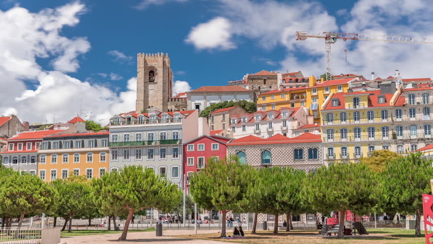 Square largo Jose Saramago timelapse hyperlapse at medieval quarter Alfama in Lisbon, sunny day with green trees and blue cloudy sky. Many colorful houses and Se de Lisboa cathedral on top. Portugal