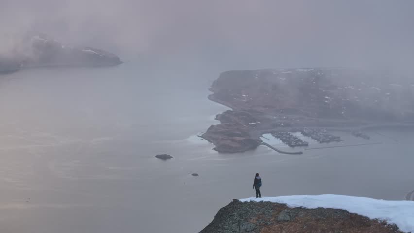 Aerial view of a majestic pyramid and a person hiking in a serene winter landscape with rugged mountains and a tranquil ocean, Unalaska, United States.