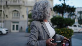 Senior businesswoman in gray suit uses mobile app to order taxi cab and raises her hand while waiting on busy city street. Female entrepreneur exudes professionalism and efficiency in metropolis - Powered by Shutterstock - Get 15% off with code: PIKWIZARD15