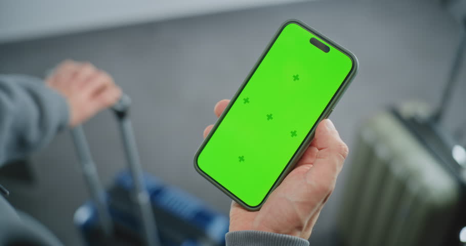 Airport Terminal: Close Up of Man Standing with Suitcase, Holding Smartphone with Green Chroma Key Display Mock Up, Going on Vacation. Video Template with Placeholder for Business Advertising Content.