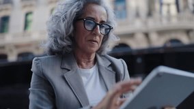 Professional businesswoman with gray hair and stylish glasses uses touch pad while discussing documents on a call. multitasking, productivity, and modern communication in an urban environment - Powered by Shutterstock - Get 15% off with code: PIKWIZARD15