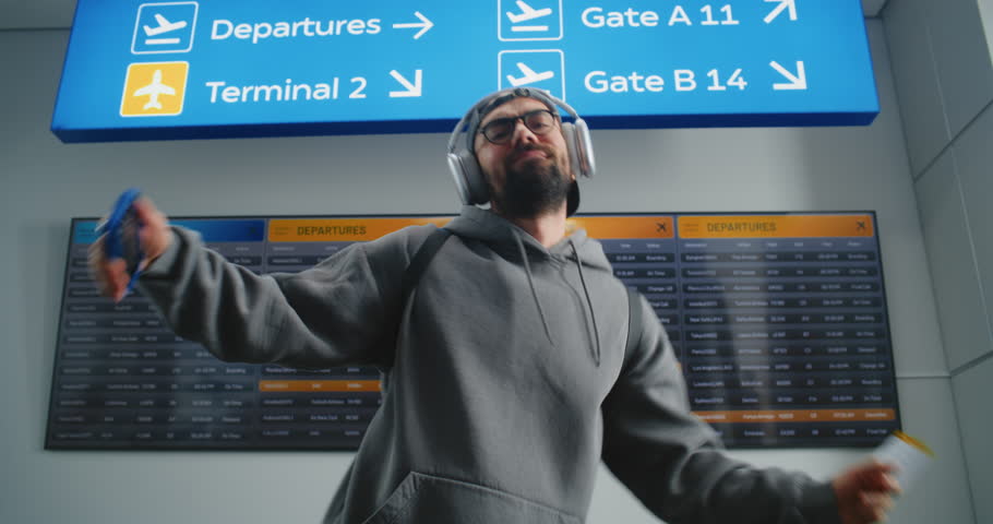 International Airport Terminal: Portrait of Happy Man Dancing, Holding Plane Ticket, Looking at Camera. Arrival and Departure Information Display in the Background. Tourist Going on Vacation Trip.